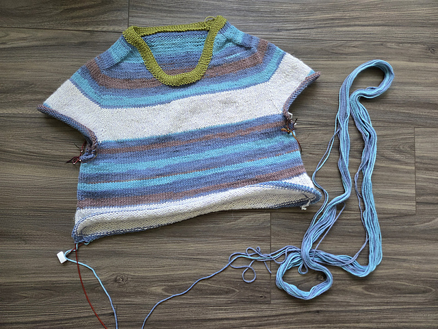 A photograph of a white and blue striped shirt being knitted in the round. It is at the stage where the body is being worked, the sleeves are unfinished, but a green collar has been added.